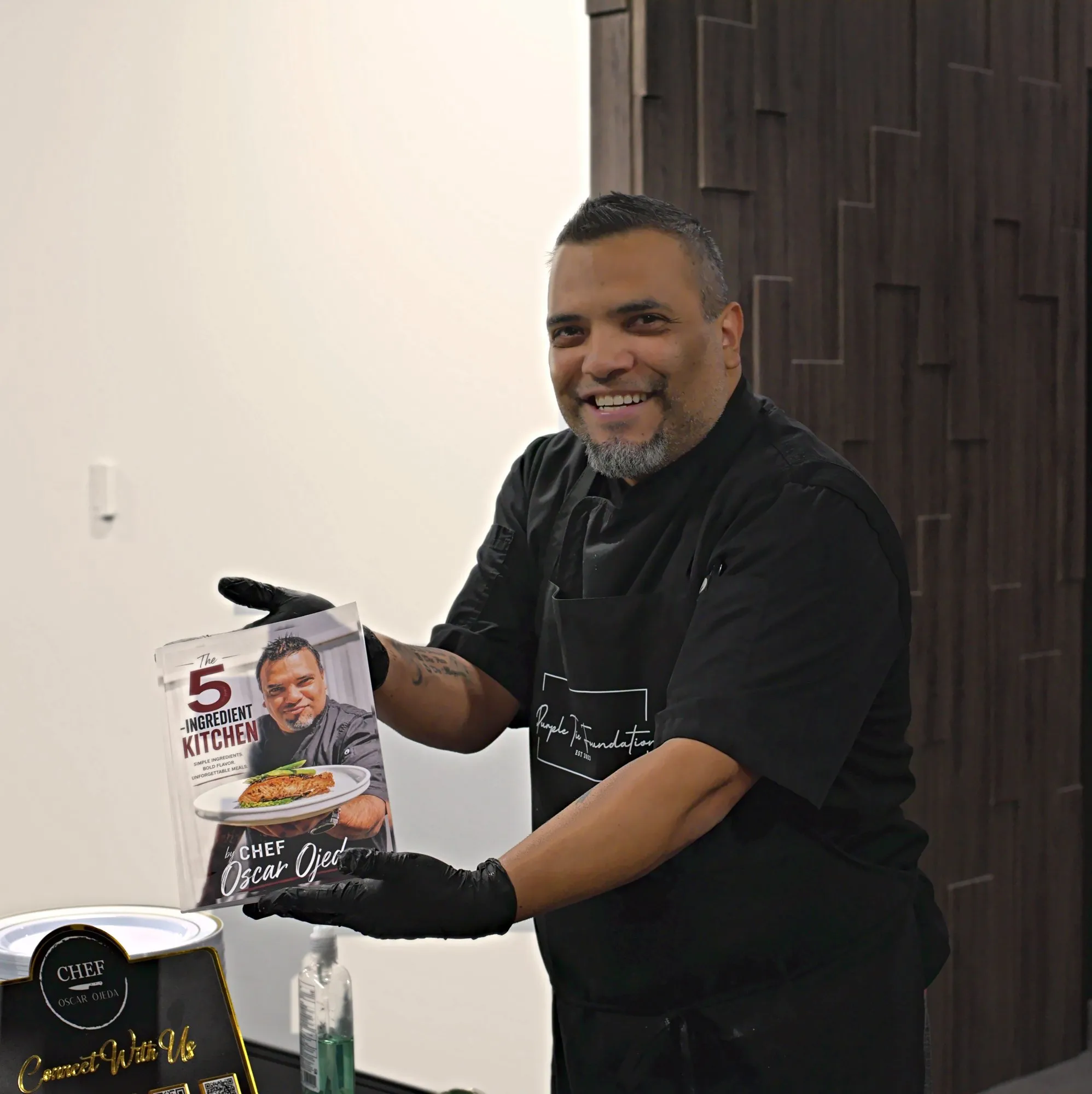 Chef Oscar Ojeda in black chef whites holding up his cookbook with a wide grin