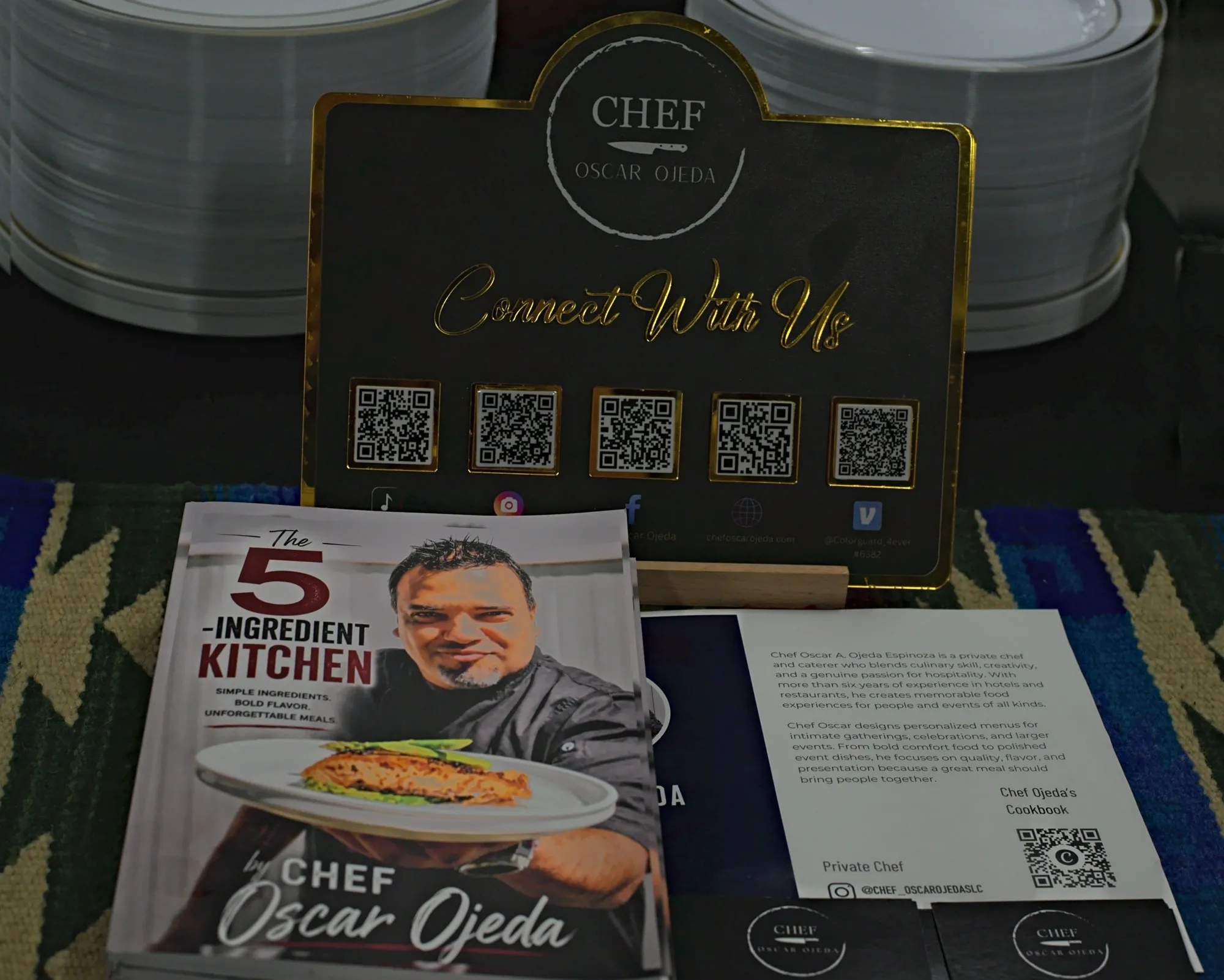 A Connect With Us sign with QR codes beside a stack of the chef's cookbook on a woven runner