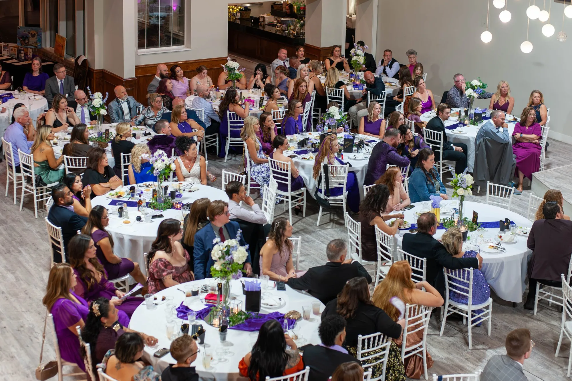 Full dining room from above, round tables dressed in white and purple, guests finding their seats