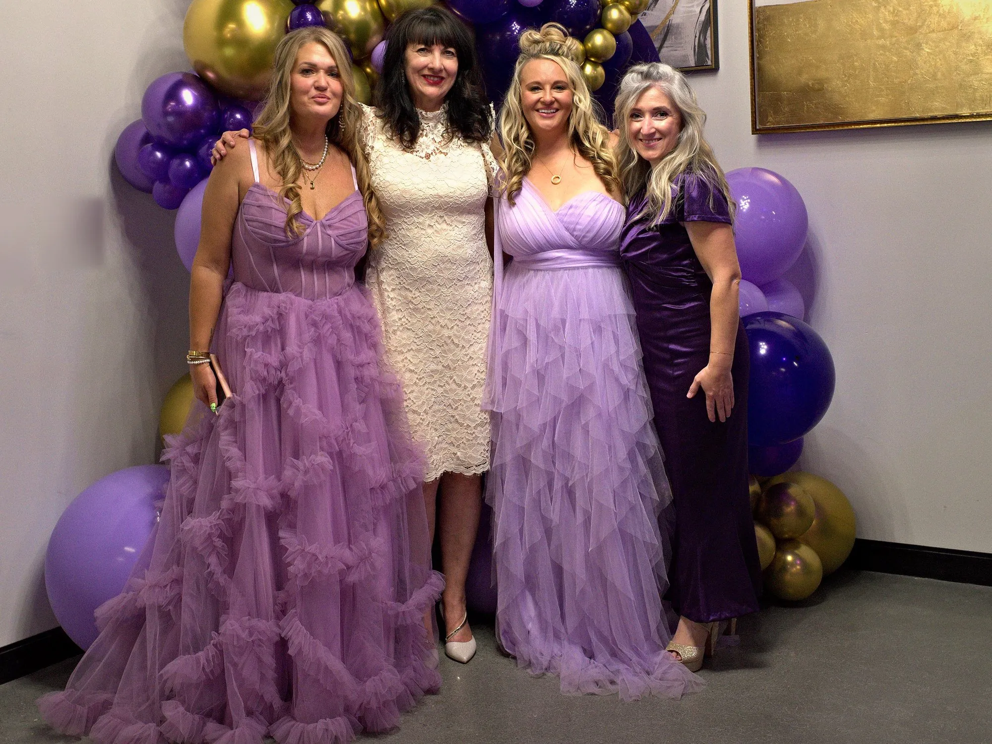 Four women in purple gowns, the foundation board, in front of a purple and gold balloon arch