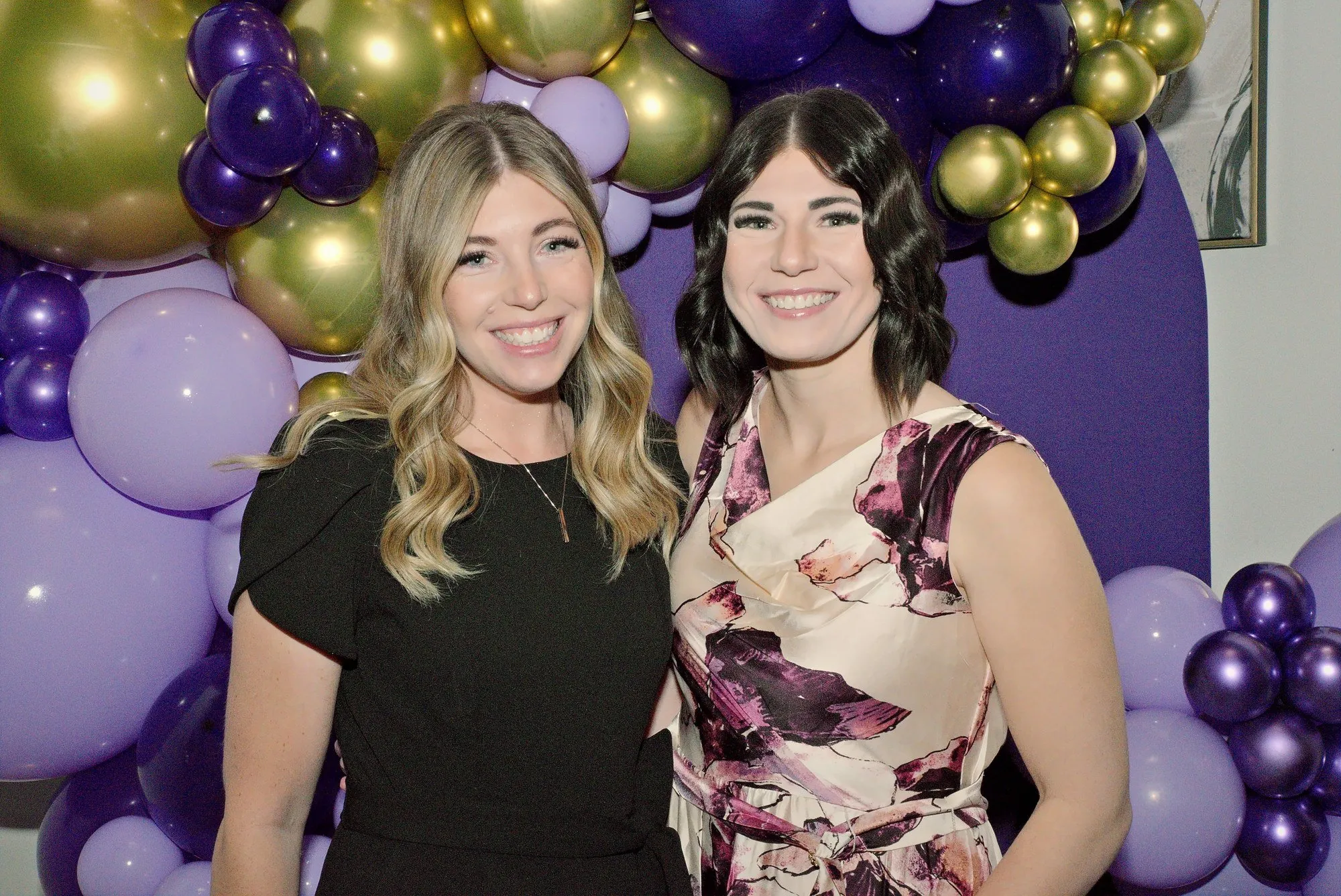 Two guests smiling at a balloon arch photo wall in purple and gold