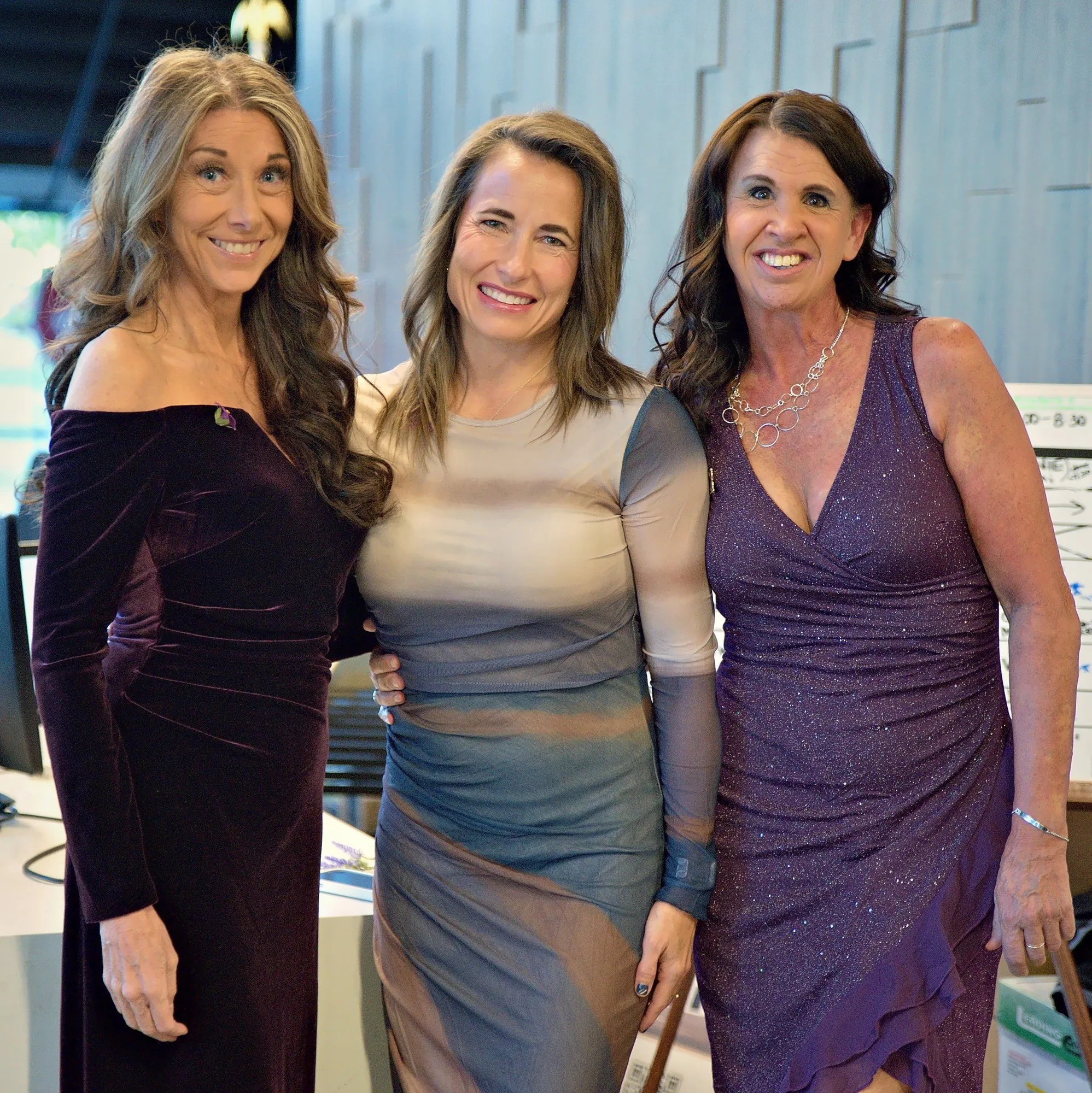 Three friends in evening dresses, arms around each other, in soft window light