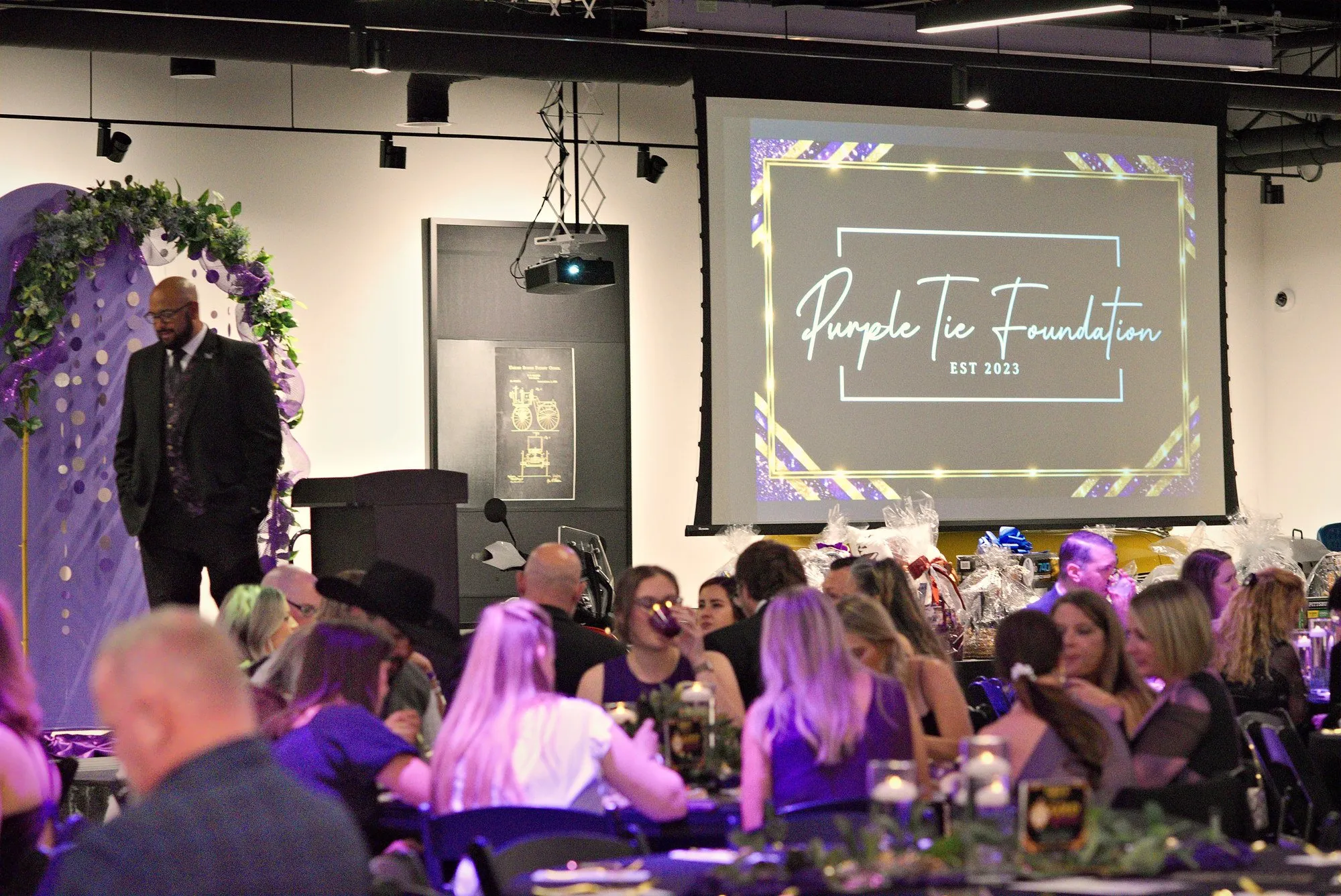 The emcee at the podium under a floral arch as the gala room watches