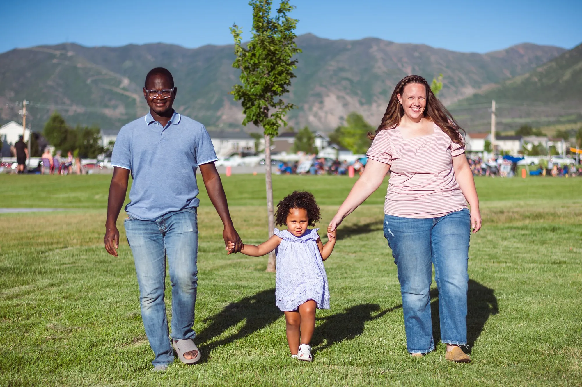 Parents walking hand in hand with their toddler across a park lawn with mountains behind them