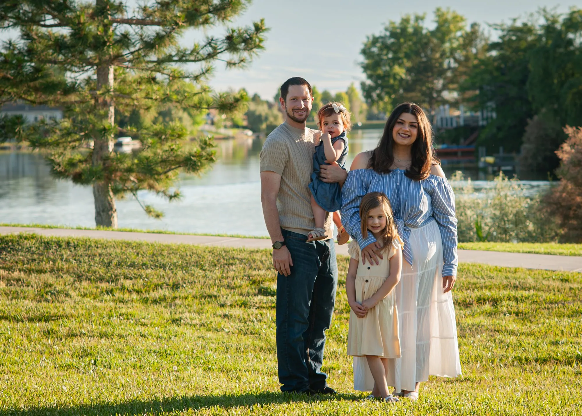 Family of four standing together on the grass beside a lake in evening light