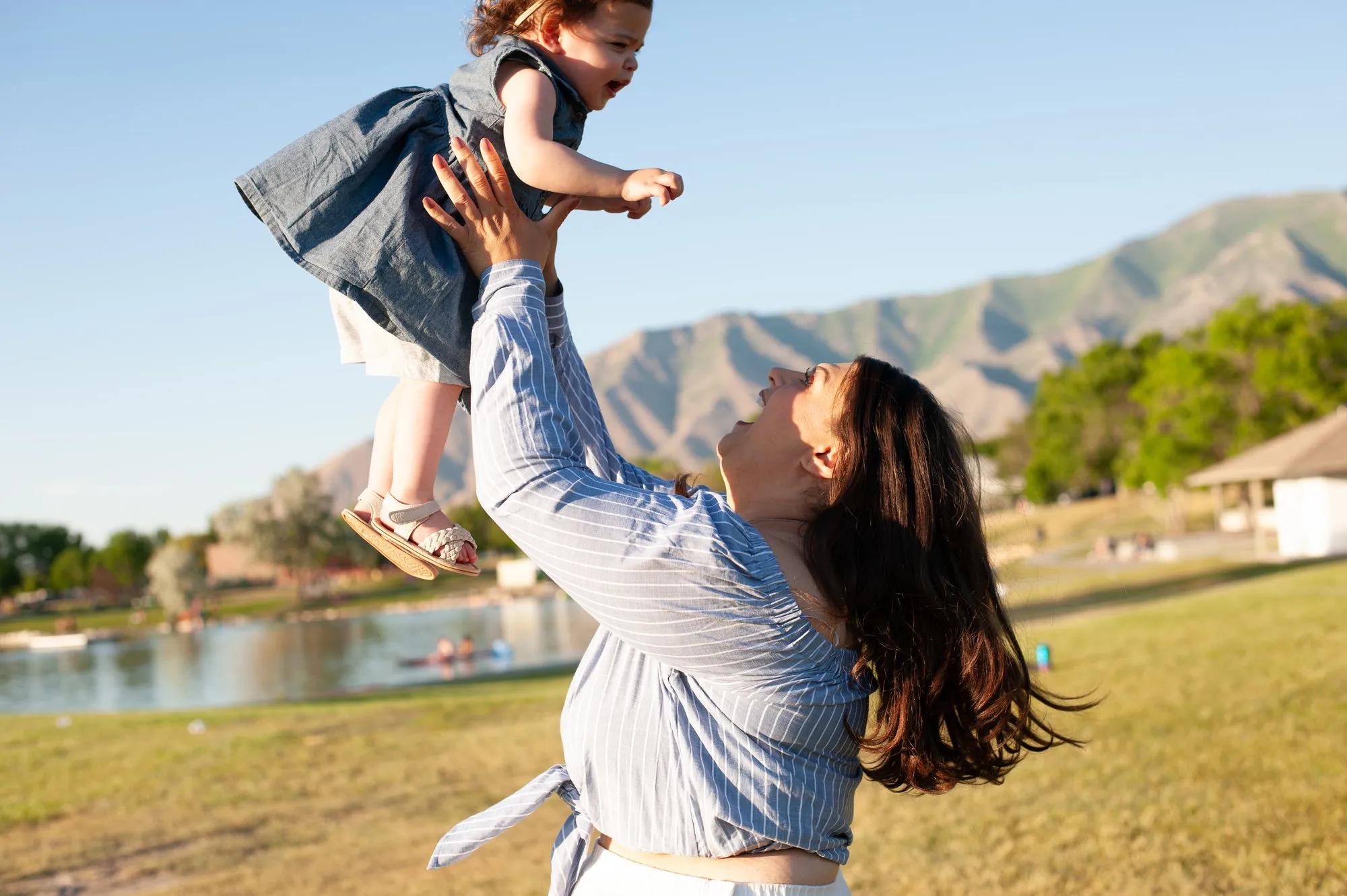 Mother lifting her toddler high in the air on a lakeside lawn