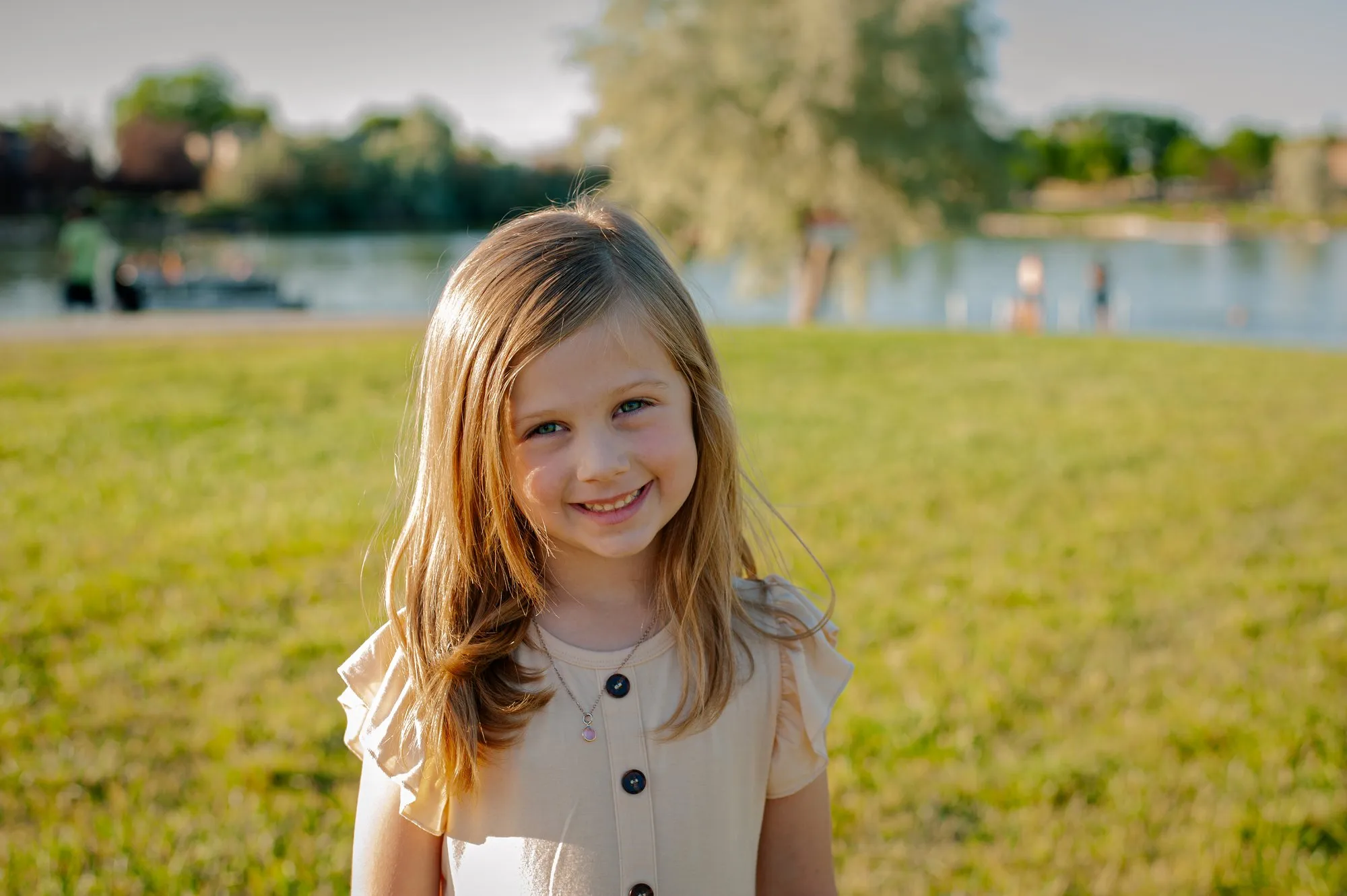 Young girl in a cream dress smiling on the grass by a lake