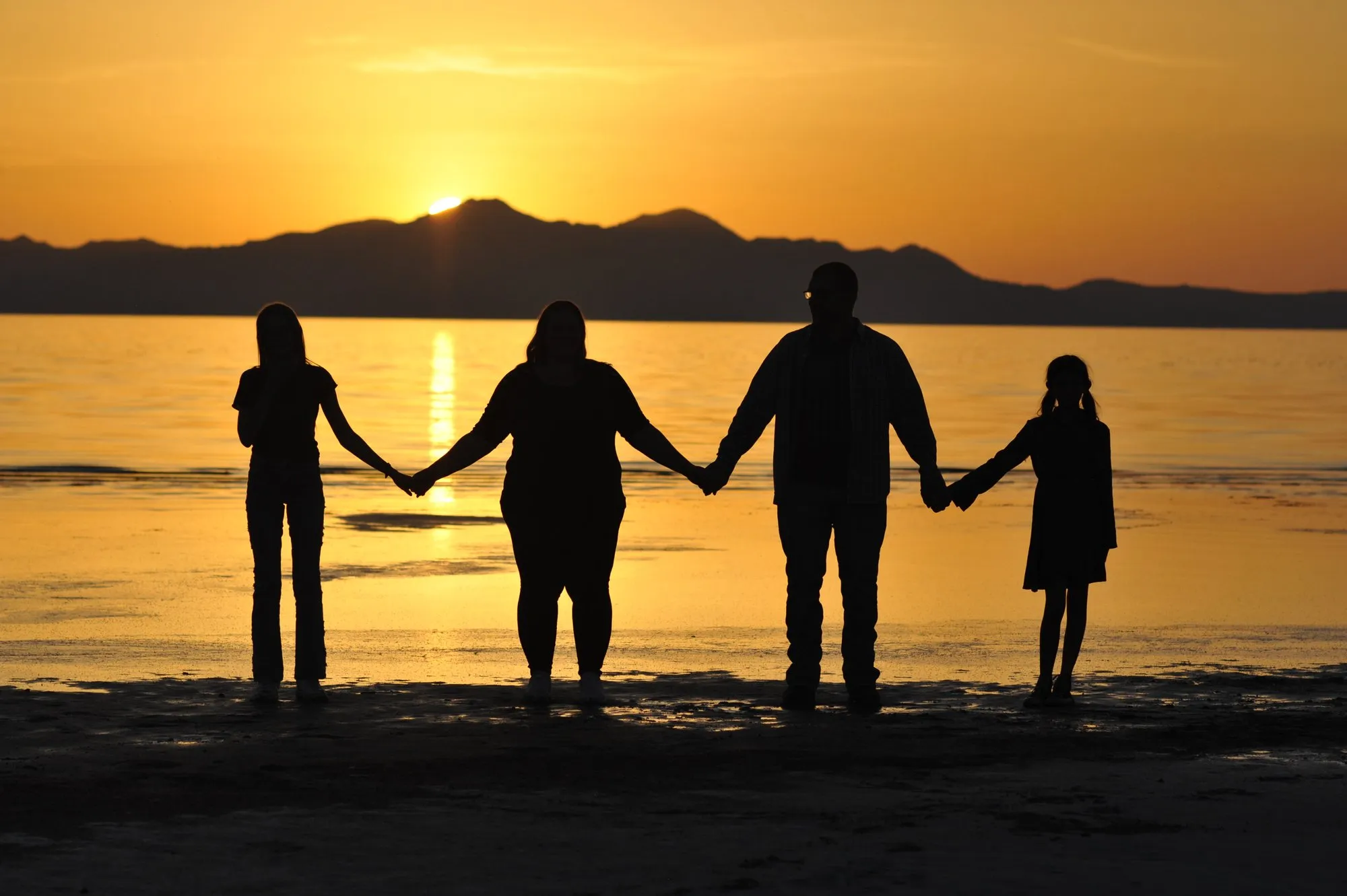 A family of four holding hands in silhouette at the edge of the Great Salt Lake as the sun sets behind the island
